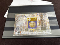 A collectible Vatican postage stamp souvenir sheet displayed inside a clear protective plastic sleeve on a striped gray holder, photographed on a dark brown textured surface.