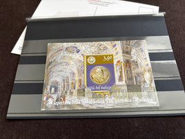 A collectible Vatican postage stamp souvenir sheet displayed inside a clear protective plastic sleeve on a striped gray holder, photographed on a dark brown textured surface.