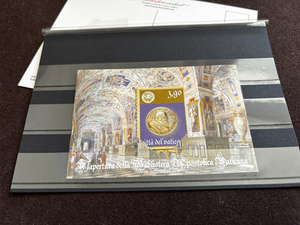 A collectible Vatican postage stamp souvenir sheet displayed inside a clear protective plastic sleeve on a striped gray holder, photographed on a dark brown textured surface.