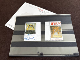 Two Vatican City postage stamps displayed side-by-side on a clear plastic sleeve over a gray striped backing card, with a white paper insert in the background.