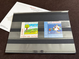 Two vintage Vatican City postage stamps displayed side-by-side on a clear plastic sleeve over a gray striped card, with a white envelope in the background on a dark brown textured surface.