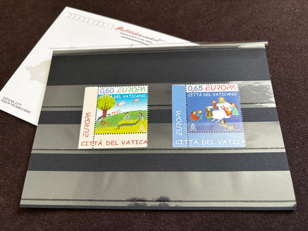 Two vintage Vatican City postage stamps displayed side-by-side on a clear plastic sleeve over a gray striped card, with a white envelope in the background on a dark brown textured surface.