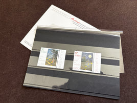 Two postage stamps displayed on a clear plastic sleeve over a striped black and gray card, with a white stamp envelope behind them, photographed on a dark brown textured surface.