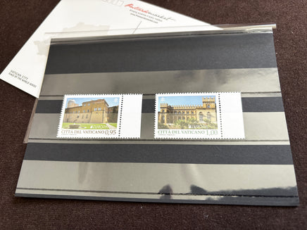 Two Vatican City postage stamps displayed side by side on a black-and-white striped protective sleeve, with part of a white envelope visible at the top left on a dark brown surface.