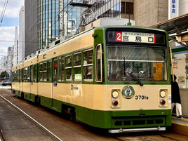 Green and beige tram on tracks in an urban setting with buildings in the background in Japan