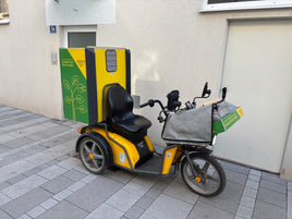 Yellow and black electric tricycle with a green box attached, parked on a sidewalk.
