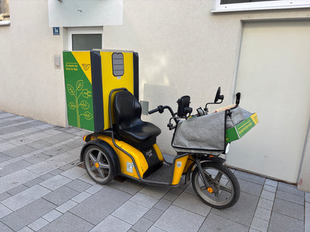 Yellow and black electric tricycle with a green box attached, parked on a sidewalk.